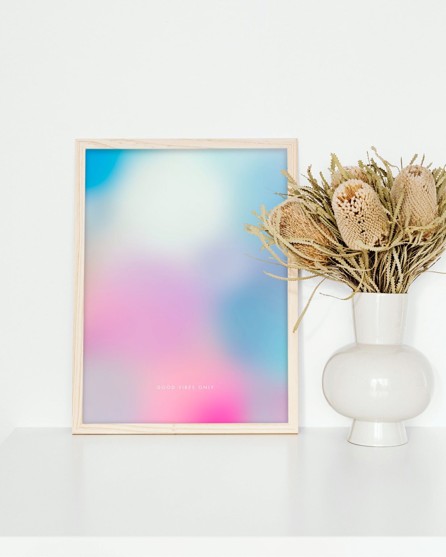 Designposter „Good Vibes Only“ eingerahmt auf weißem Regal neben weißer Vase und Trockenblumen, modernes Interior.