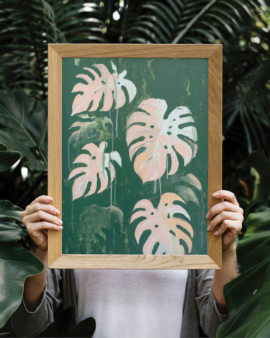 Ein Gemälde mit Monstera-Blättern in einem Holzrahmen wird von einer Person hochgehalten. Ihr Gesicht ist dadurch verdeckt. Im HIntergrund sieht man Palmblätter