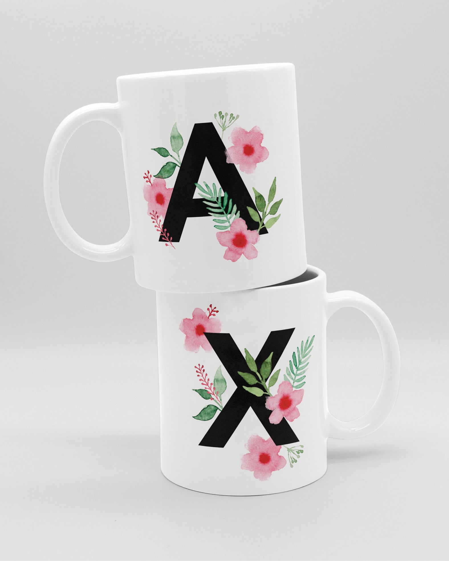 Zwei weiße Keramiktasse aufeinander,  mit schwarzem Buchstaben A und X und rosa-grünen Aquarell-Blumen