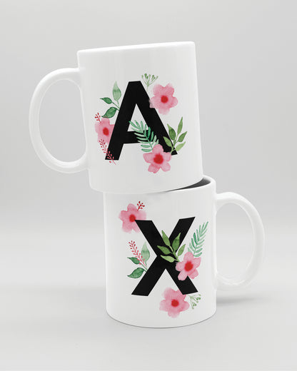 Zwei weiße Keramiktasse aufeinander,  mit schwarzem Buchstaben A und X und rosa-grünen Aquarell-Blumen