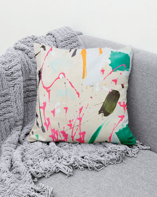 Beiges Kissen mit bunten Farbklecksen auf einem grauen Sofa.