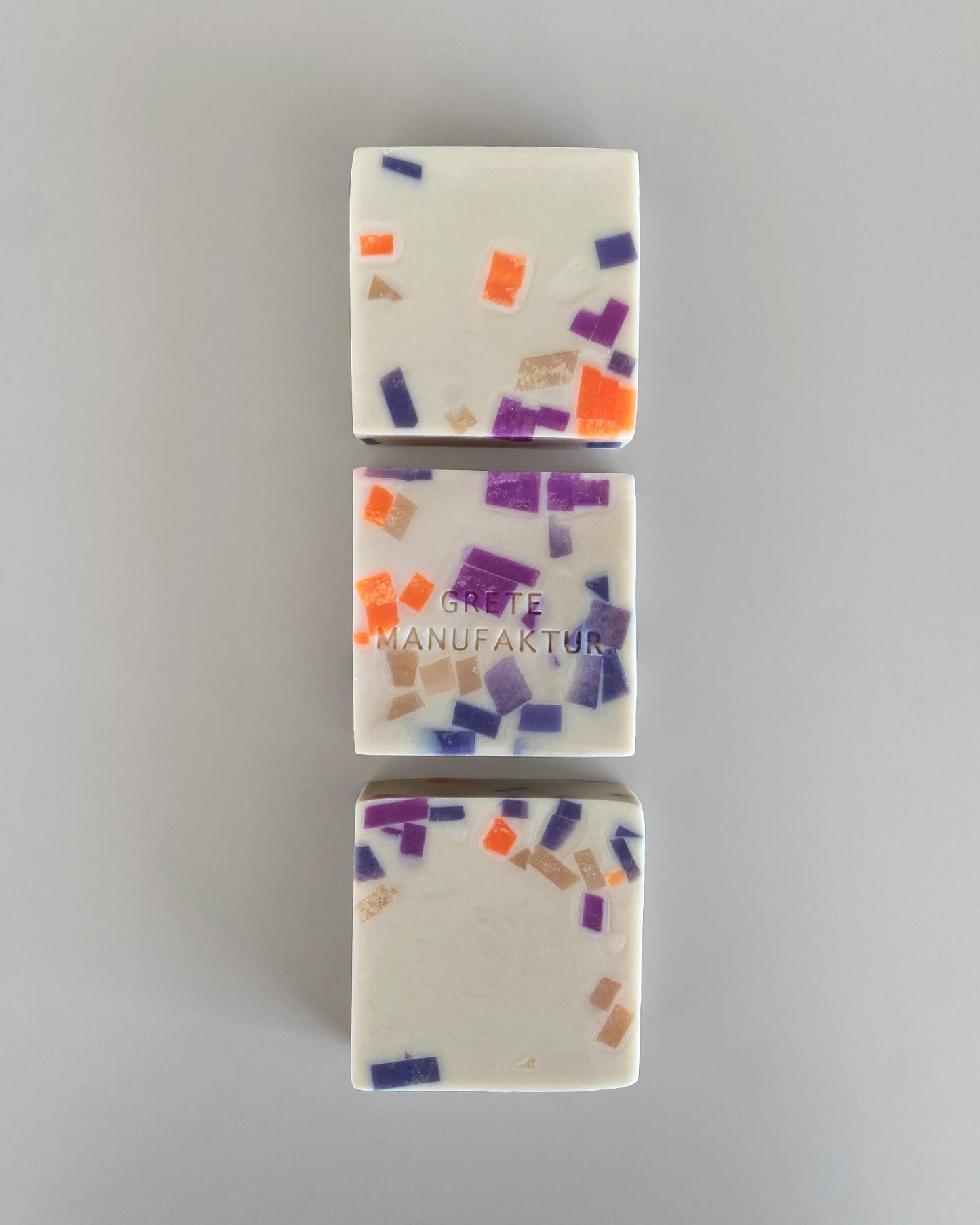 Drei quadratische Seifenstücke gerade untereinander gelegt auf grauem Hintergund. Die Seifen sind Off-White, sie haben ein Terrazzo-Muster aus neonorangenen, lilanen und nudefarbenen Stücken. Sie mittlere Seife hat die Worte Grete Manufaktur eingeprägt