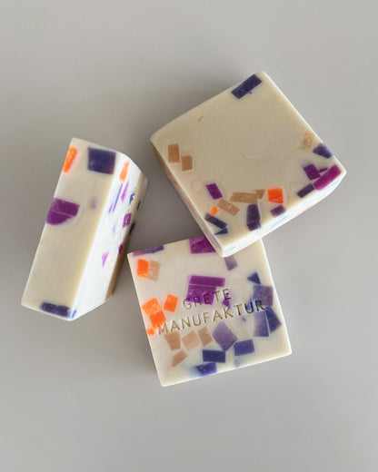 Drei quadratische Seifenstücke sind locker gelegt auf einem grauen Hintergrund. Das Foto wirkt inszeniert und hochwertig. Die Seifenstücke haben ein besonderes Design: die Grundfarbe ist Off-White, durchzogen mit kleinen Seifenstücken in Neonorange, Lila und Nude ergibt sich daraus eine Terrazzo Ästhetik.