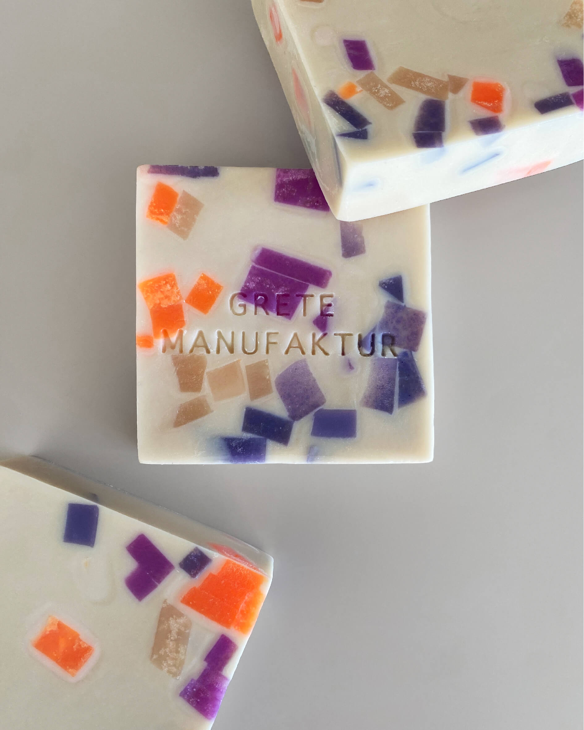 Drei cremefarbene, quadratische Seifenstücke mit Sprenkeln ist Neonorange, Lila und Nude liegen arrangiert auf einem grauen Hintergrund