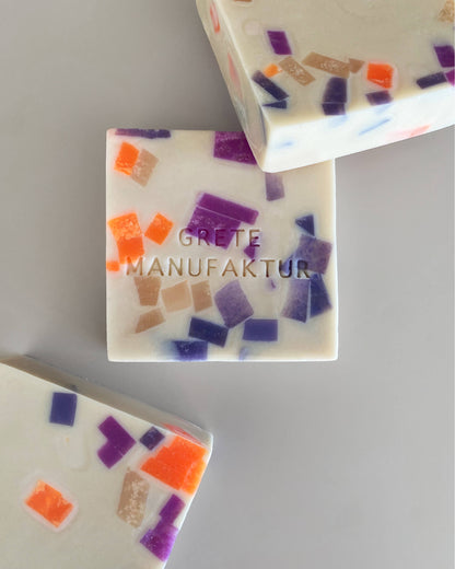 Drei cremefarbene, quadratische Seifenstücke mit Sprenkeln ist Neonorange, Lila und Nude liegen arrangiert auf einem grauen Hintergrund