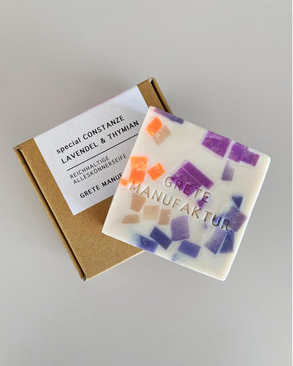Seife in Off-White mit Terrazzo-Optik mit Neon-Orange, Lila und Nude auf einer Verpackung aus Pappe, mit Text auf Aufkleber: special Constanze Lavendel & Thymian, reichahltige Alleskönnerseife, Grete Manufaktur