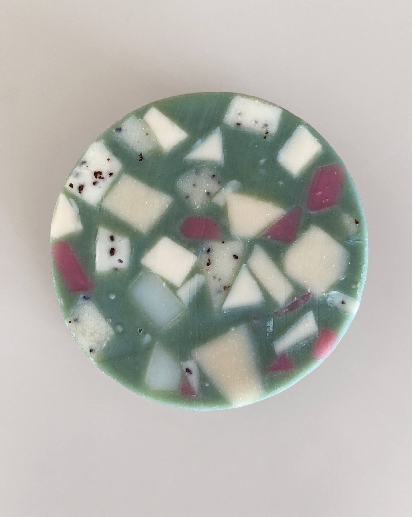 Eine natürlich und zugleich hochwertig wirkende runde Seife liegt auf einem grauen Hintergrund. Die Grundfarbe ist ein dunkler Minze-/Türkis-Ton der mit geometrischen Fragmenten in Weiß und Lila durchzogen ist. Man sieht Spuren des handgemachten Herstellungsprozesses. 
