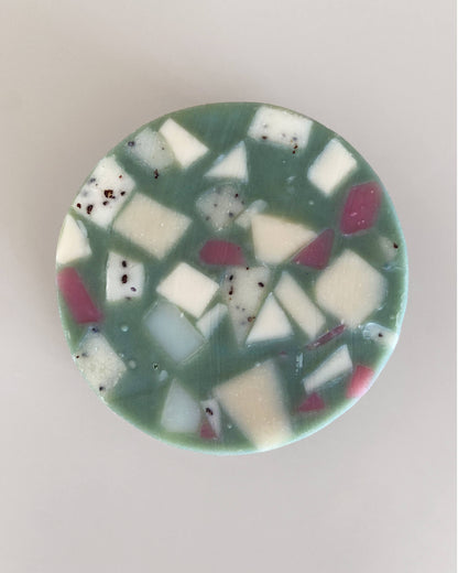 Eine natürlich und zugleich hochwertig wirkende runde Seife liegt auf einem grauen Hintergrund. Die Grundfarbe ist ein dunkler Minze-/Türkis-Ton der mit geometrischen Fragmenten in Weiß und Lila durchzogen ist. Man sieht Spuren des handgemachten Herstellungsprozesses. 