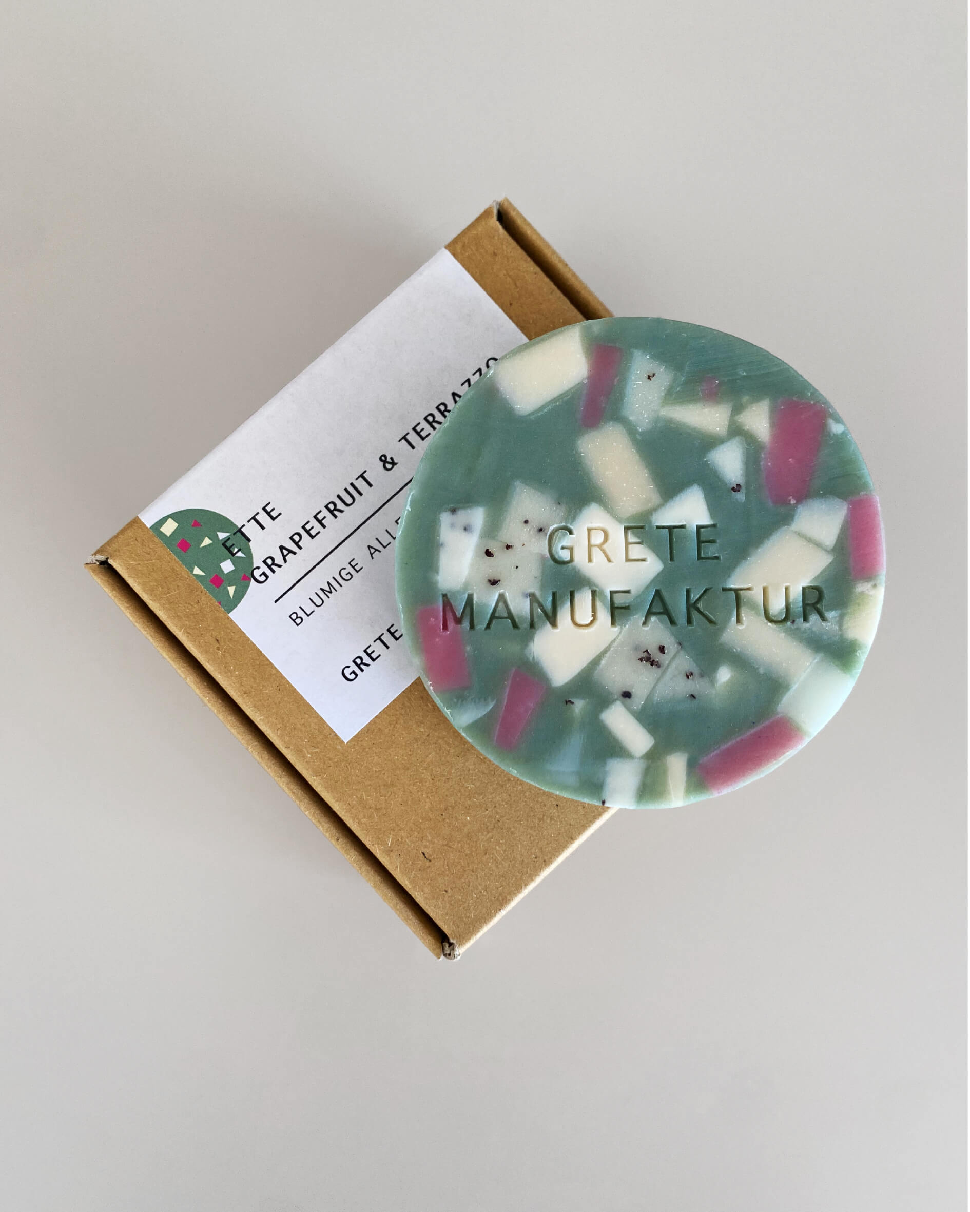 Ette Seifen im Terrazzo-Design in Türkis mit geometrischen Fragmenten in Weiß und Lila. Grete Manufaktur ist in die Oberfläche geprägt. Die Seife liegt auf einer Pappschachtel mit einem beschrifteten Aufkleber. Text: Ette Grapefruit & Terrazzo, Blumige alleskönnerin, Grete Manufaktur.