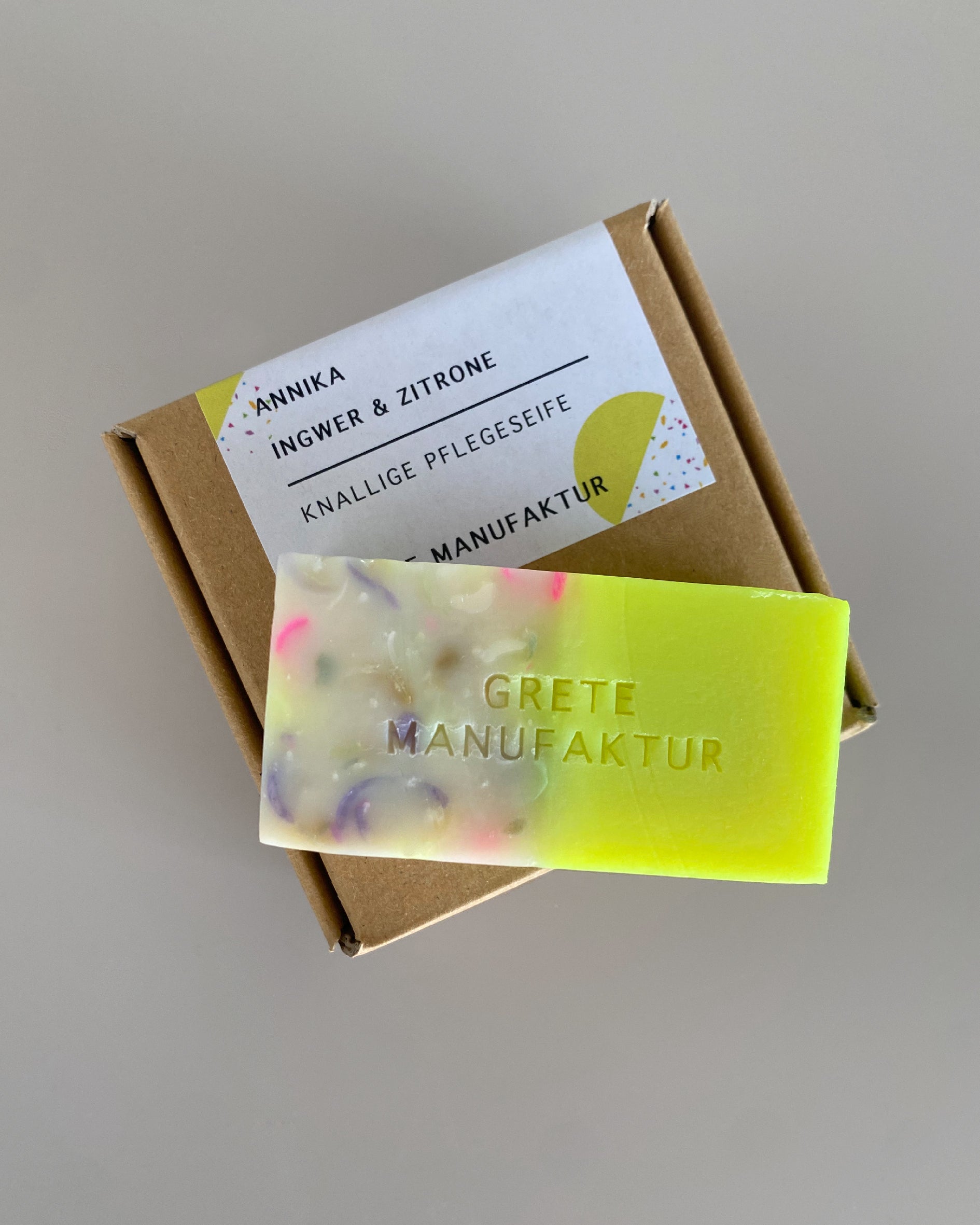 Eine Seife liegt auf einer Verpackung aus Naturpappe vor grauem Hintergrund. Die Seife ist auf einer Seite bunt, auf der anderen Neongelb.