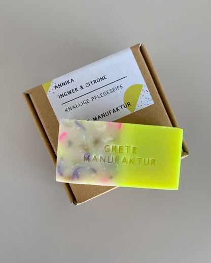 Eine Seife liegt auf einer Verpackung aus Naturpappe vor grauem Hintergrund. Die Seife ist auf einer Seite bunt, auf der anderen Neongelb.