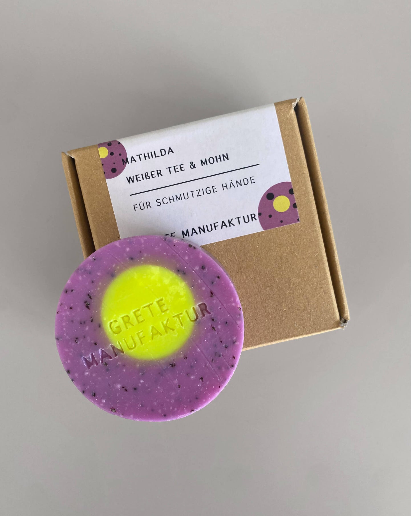 Eine lila Seife mit neongelbem rundem Dekor liegt auf einer Schachtel aus Pappe mit beschriftetem weißem Aufkleber. Text Mathilda, weißer Tee & Mohn für schmutzige Hände von Grete Manufaktur